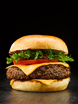 Food photograph of a cheeseburger, a golden brioche bun topped with a juicy seared homemade burger patty, thick sliced fresh tomato, lettuce and two slices of Ultimate BBQ Melts cheese, shot on a black background with a pack of Ultimate BBQ Melts overlayed on the image, showing the burger on the front of the pack