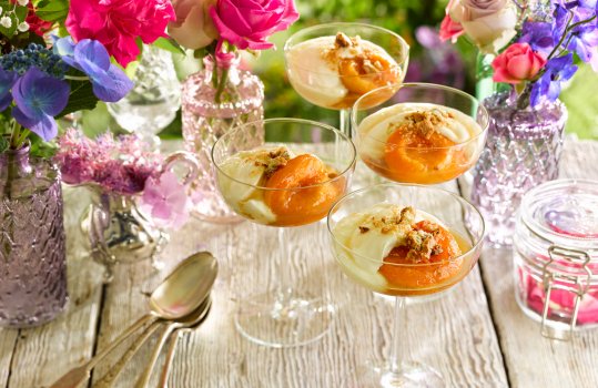 Food photograph of poached shiny glazed apricots served with zabaglione and chopped mixed nuts in champagne coups on an outdoor picnic table surrounded by small glass vases of flowers, with foliage in the background