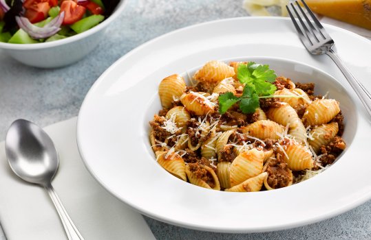 Food photograph of a bowl of conchiglie bolognese, rich bolognese sauce coating conchiglie pasta and topped with shaved parmesan, shot on a grey stone background alongside a bowl of fresh salad and a block of parmesan cheese