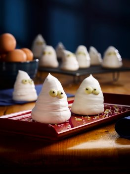 Food photograph of Hallowe'en ghost meringues, meringues in the shape of ghosts with chocolate eyes stuck on them, served on a red rectangular plate on a dark wooden tabletop with a bowl of eggs and more ghost meringues on a cooling rack in the background