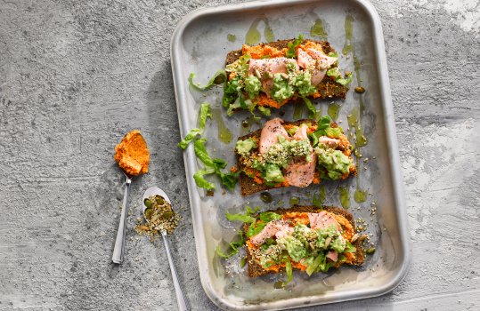 Aerial food photograph of flaked salmon with avocado, shredded lettuce, mixed seeds and kimchi butter on pumpernickel bread, drizzled with olive oil and shot on a steel baking tray on a grey stone background