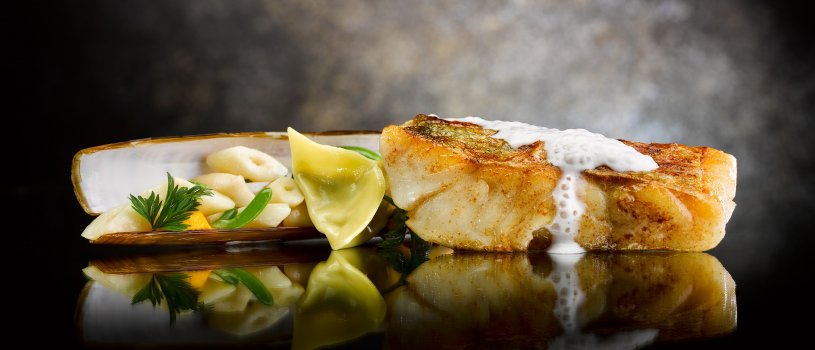Food photograph of a fine dining fish course, a fillet of pollock with crispy golden skin topped with cream sauce, served with a steamed razor clam served in its shell, with sea vegetables and a clam tortellini. Shot on a reflective mirrored black and grey background