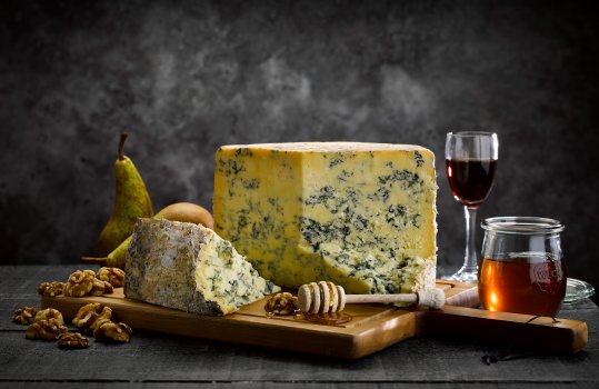 Food photograph of a half wheel of crumbly blue stilton cheese, shot on a dark set on a wooden board with honey, walnuts, pears and a glass of port in the background