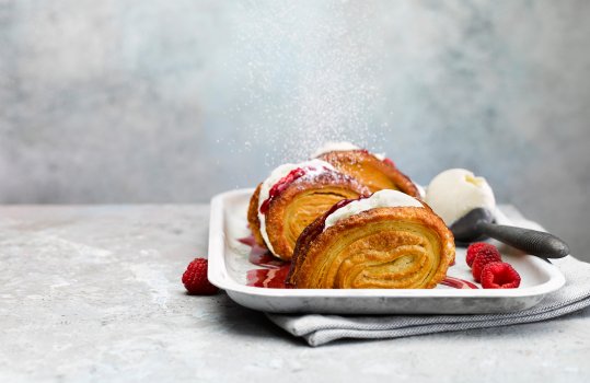 Food photograph of a palmier ice cream sandwich, a golden flaky palmier cut in half and filled with raspberry jam and vanilla ice cream, and dusted with icing sugar. Shot on a steel baking tray alongside fresh raspberries and a vintage ice cream scoop on an abstract light grey set