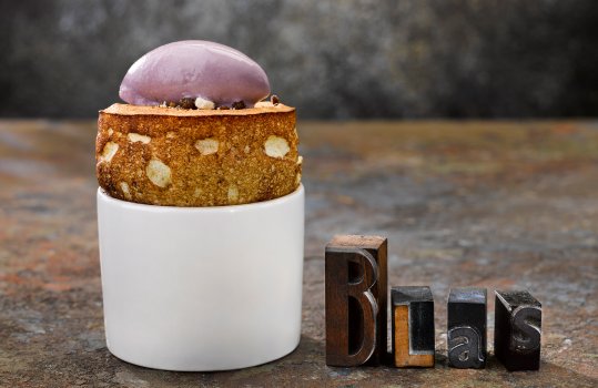 Food photograph of a fine dining dessert, a risen golden brown blackberry souffle topped with a rocher of parma violet ice cream, shot on an abstract painted background with antique letterpress spelling out the word Blas, the Welsh word for taste.