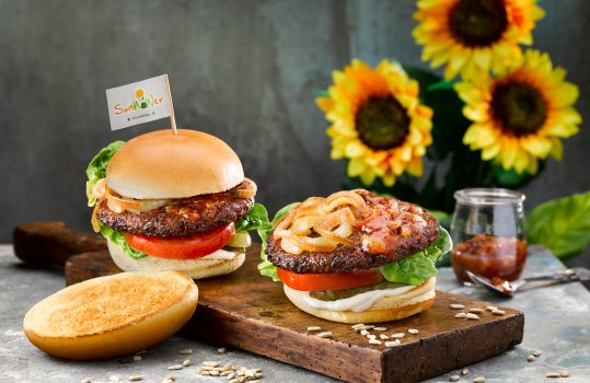 Food photograph of two SunWOWer burgers, vegan burgers made from sunflower seeds served on vegan brioche style buns with caramelised onions, thick sliced fresh tomato, lettuce, pickles and mayonnaise. Shot on a wooden board on a grey background with a glass jar of chutney and sunflowers