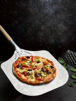 Food photograph of a vegetarian tomato, artichoke, olive and pepper pizza shot on a dark grey background with fresh basil leaves and an antique cheese grater, on a steel pizza peel