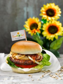 Food photograph of a SunWOWer burger, vegan burger made from sunflower seeds served on vegan brioche style buns with caramelised onions, thick sliced fresh tomato, lettuce, pickles and mayonnaise. Shot on parchment paper on a grey background with a glass jar of chutney and sunflowers
