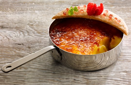 Food photograph of a crispy shiny caramel topped creme brulee, served in an individual steel pan, alongside a crisp raspberry shortbread, served on a grey wooden table