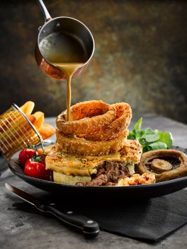Food photograph of a pub lunch, steak pie with golden crisp pastry topped with panko breaded onion rings and served with roasted tomatoes, mushroom and chips, with peppercorn sauce being poured over from a copper pot. Shot on a dark grey background