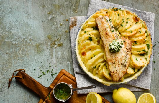 Aerial food photograph of an oven baked haddock fillet sitting atop an enamel dish of pommes Anna, sliced potatoes cooked in butter and cream, shot on a blue grey background with a pot of garlic butter and lemon halves