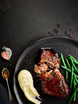 Four aerial food photographs that feature on the cover of frozen meat products, confit duck legs with gravy, beef bourgignon, glazed lamb shoulder and buffalo chicken wings, all shot on black plates and bowls on a black slate background
