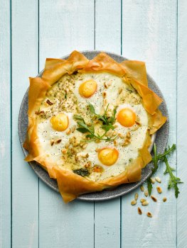 Aerial food photograph of a breakfast filo pastry pie, crispy layers of filo pastry filled with a spinach and pine nut filling, with eggs cracked on top before being baked, served on a grey plate on a pale blue wooden background