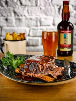 Food photograph of a grill restaurant main course, a braised, roasted and glazed beef short rib which is shredding off the bone, served with a cup of skin on thick cut chips and a pint of beer, on a wooden table with a white painted brick background
