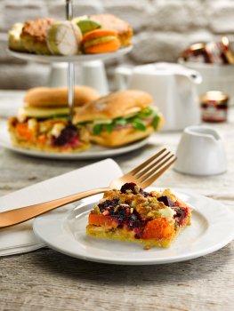 Food photograph of a beetroot and butternut squash slice, roasted and topped with pine nuts, served on a white plate in an afternoon tea setting with sandwiches and macarons in the background next to a teapot and a milk jug, shot in a grey wood setting