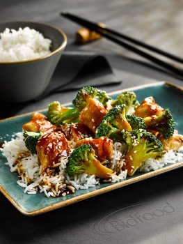 Food photograph of a chicken and broccoli stir fry, shiny glazed chunks of chicken and broccoli florets in a sweet and sour sauce, served over steamed white rice on a teal plate, with a bowl of rice and chopsticks in the background, served on a leather mat on a grey tabletop