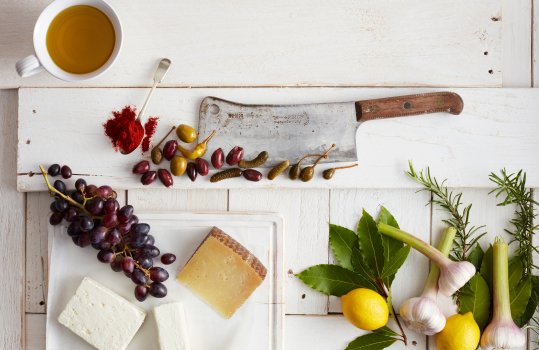 Aerial food photograph of the ingredients to make Spanish coca bread shot on painted white wooden planks, featuring feta, manchego and goats cheeses with grapes, olives, pickles, capters, fresh lemon, fresh bay and rosemary leaves, and fresh garlic bulbs