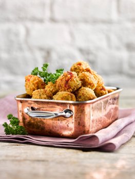 Food photograph of deep fried red pepper risotto bites, breaded balls of risotto with a crispy golden exterior served in a copper dish on top of a lilac napkin garnished with curly parsley, shot on a grey wooden table with white painted bricks in the background