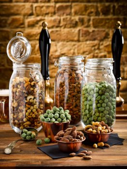 Food photograph of various bar snacks, mixed nuts, wasabi peas and smoked almonds, in large kilner jars with portions in copper ramekins; on a wooden tabletop with a brick wall, a glass of beer and beer pumps in the background