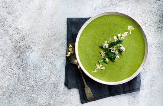 Aerial food photograph of a bowl of homemade broccoli and stilton soup, garnished with small broccoli florets and crumbled stilton cheese. Served in a white ceramic bowl on a navy blue napkin, on a light grey tabletop