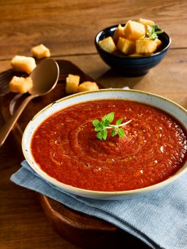 Food photograph of a homemade tomato soup served in a shallow bowl drizzles with olive oil and topped with Greek basil, served with a bowl of golden croutons on a dark wooden background
