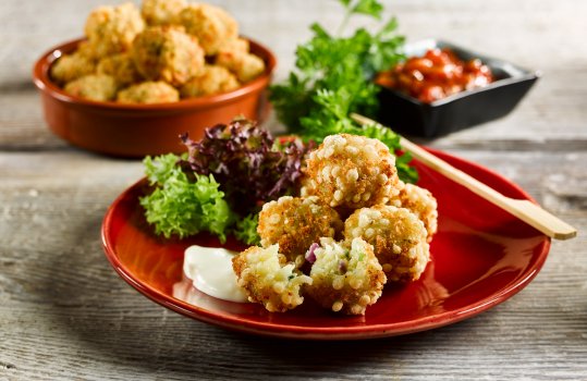 Food photograph of deep fried red pepper risotto bites, breaded balls of risotto with a crispy golden exterior served on a red ceramic dish garnished with mixed leaves and curly parsley, shot on a grey wooden table
