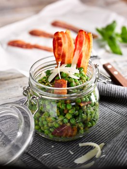 Food photograph of a pea, pesto and bacon mix served in a glass kilner jar topped with shaved parmesan cheese and two slices of crispy streaky bacon. Shot in a light grey setting with a blue fabric napkin