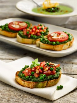 Food photograph of a pea and mint bruchetta, golden toasted slices of baguette topped with a crushed pea, mint and pesto mix, along with diced tomato, garlic and pine nuts. Served on a white napkin with a plate of bruschetta in the background, and a bowl of pea and parmesan soup, all on a grey tabletop