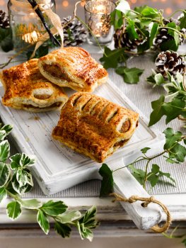 Food photograph of a festive sausage roll, crisp golden flaky pastry surrounding a sausagemeat filling shot on a painted white chopping board on a vintage white sideboard, with candles, fairy lights, ivy and pinecones