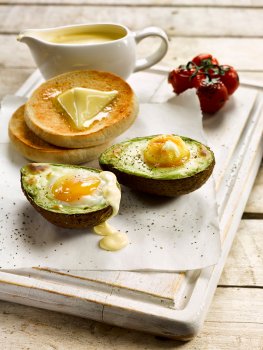 Food photograph of a brunch dish of avocado stuffed with eggs and baked, served on a white painted wooden board alongside hot toasted buttered English muffin with roasted vine cherry tomatoes and hollandaise sauce