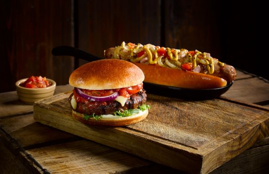 Food photograph of a juicy griddled burger patty with lettuce, onion, cheese and salsa inside a golden brioche bun, and a loaded hot dog topped with mustard, pickles, caramelised onions and ketchup sitting on a split top brioche bun in a cast iron tray. Shot on a dark wooden set with a wooden board, a vintage wooden apple crate and a dark wooden background