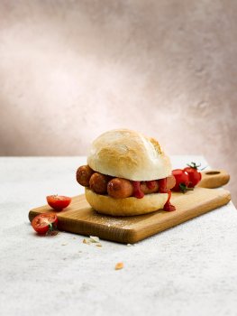Food photograph of a vegan sausage sandwich, three dark brown pan fried sausages inside a crusty white bread roll with tomato ketchup, served on a wooden board with vine cherry tomatoes in an abstract warm grey set