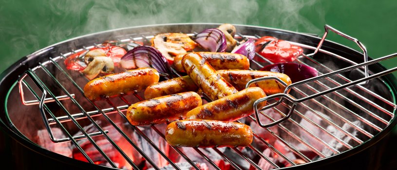 Food photograph of eight golden bar mark charred chicken sausages alongside red onion, red pepper and mushrooms shot on a lit barbecue in front of a green background with billowing smoke coming from the barbecue and a pair of barbecue tongs lifting one sausage