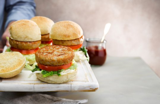 Food photograph of a wooden board with home cooked family style chicken burgers, rustic crusty bread rolls topped with mayonnaise, shredded lettuce, thick sliced fresh tomatoes and seared golden brown chicken burger patties, shot on an airy light grey background with a glass jar of ketchup and a mans arm holding the board of burgers