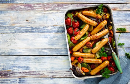 Aerial food photograph of a home style chicken sausage bake, ten thin chicken sausages baked in a steel tray amongst various vegetables including vine cherry tomatoes, leek, red onion, peppers and carrots. Shot on a painted blue and purple wooden background and sprinkled with curly parsley leaves