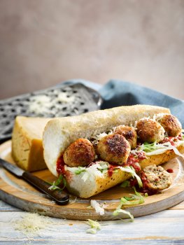Food photograph of a home style chicken meatball sub, a crusty baguette topped with tomato sauce, lettuce and seared chicken meatballs, with grated parmesan cheese scattered over the top. Shot on a light painted wooden background with a blue fabric napkin and a vintage cheese grater