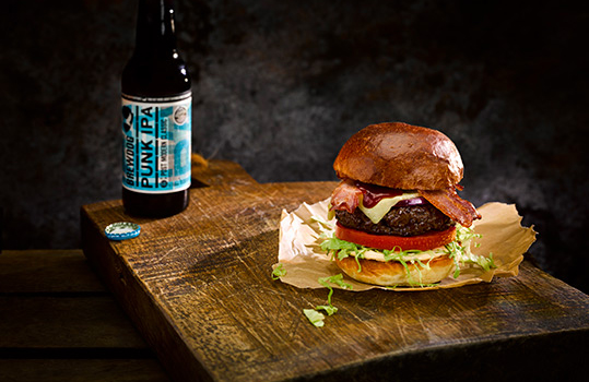 Dark moody food photograph of a homemade beef burger, a thick seared beef patty topped with melted cheese, crispy bacon and BBQ sauce, with fresh tomato and lettuce in a golden brioche bun, served on baking paper on a rustic antique chopping board with a bottle of BrewDog Punk IPA