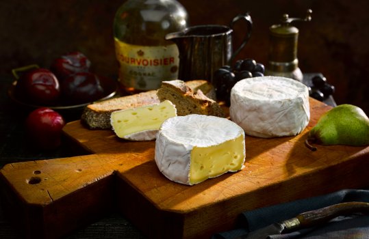 Food photograph of two wheels of brie, shot in a dark setting on a vintage chopping board alongside fresh bread, pears, plums and dates, with a vintage bottle of brandy and antique tableware in the background