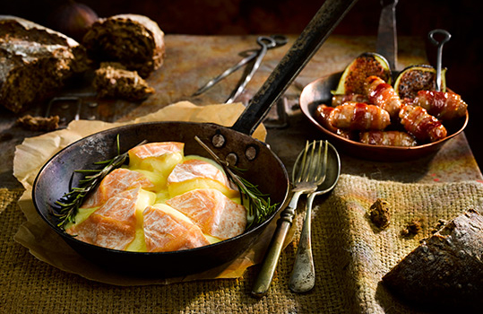 Food photograph of a cooked wheel of Golden Cenarth cheese, served with rosemary in a vintage iron frying pan alongside vintage cutlery, fresh crusty bread, golden pigs in blankets and roasted figs