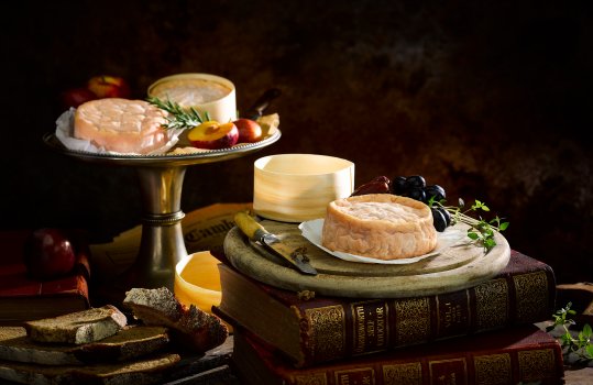 Food photograph of a wheel of Golden Cenarth cheese, shot in a dark setting on a vintage chopping board and an antique cake stand, alongside rosemary, crusty bread, plums, dates and grapes