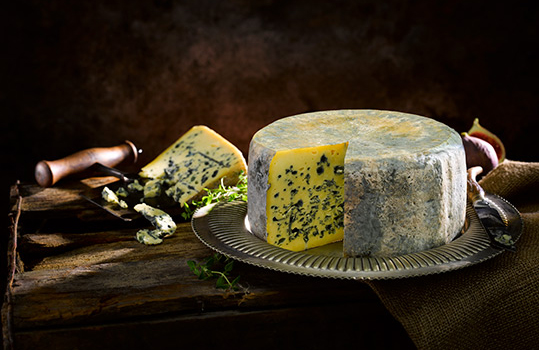 Food photograph of a whole wheel of Perl Las cheese with a mottled blue exterior and a golden inside flecked with blue veins, the whole wheel is shot on a vintage metal plate with a wedge cut from it and presented behind the wheel, crumbled slightly onto the tabletop
