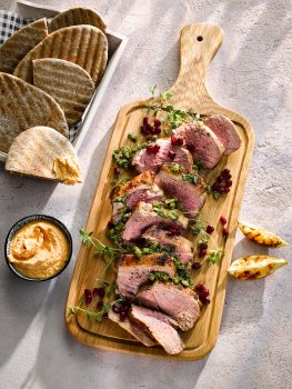 Food photograph of a juicy seared medium rare lamb leg steak, topped with a green olive tapenade and pomegranate seeds and served with houmous, grilled pita bread and charred lemon wedges. Served on a wooden board on a grey tabletop