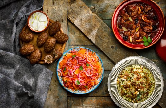 Aerial food photograph of an array of Moroccan and Middle Eastern inspired dishes, homemade lamb kibbeh with lebneh, carrot, pickled onion, mint and orange salad, chicken tagine with preserved lemon, and rice and lentil pilaf, shot on a dark weathered wooden background