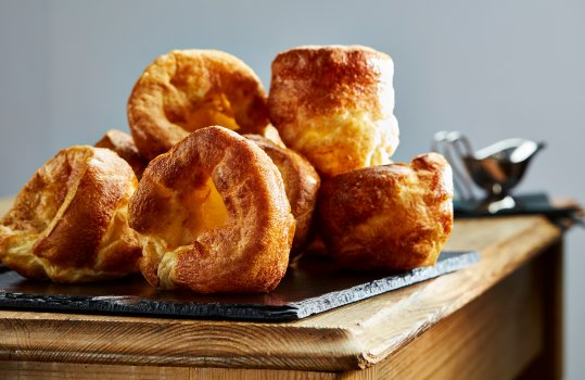 Food photograph of a pile of golden crispy Yorkshire puddings, shot on a vintage farmhouse table alongside a gravy boat