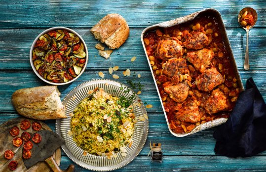 Aerial food photograph of an array of homemade Spanish inspired dishes, chicken, chorizo and white bean stew served alongside flaked almond and saffron rice; shot with orzo ratatouille, roasted tomatoes and fresh baked bread, shot on a blue painted background