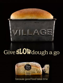 Food photograph of a loaf of bread baking in a tin branded with the word VILLAGE, with the golden top crust rising above the edge of the tin. Shown above advertising graphics alongside a photograph of a packaged sliced loaf of Village Bakery bread