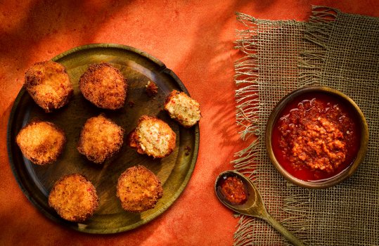 Aerial food photograph of a portion of golden crisp Akara, West African bean and red pepper fritters, served with a wooden bowl of Nigerian pepper sauce, Ata DinDin