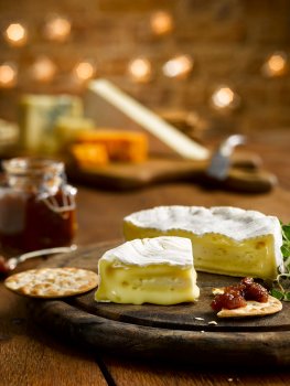 Food photograph of asoft camembert cut into wedges and served on a wooden board with crackers and chutney, with brie, stilton and cheddar on a wooden board in the background. All served on a wooden tabletop with a brick wall and Christmas lights in the background.