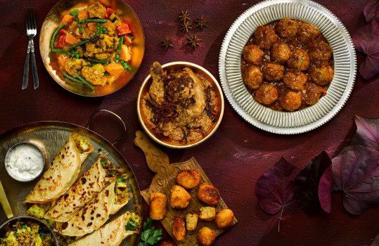 Aerial food photograph of a selection of Indian dishes, including masala dosa served on a large brass tray with raita, vegetable and chicken curries served in golden bowls, and a kofta curry served in a tin dish, all shot on a dark red background with scattered Autumn leaves