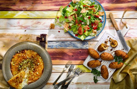 Aerial food photograph of a selection of Syrian recipes, stuffed kibbeh with lamb filling, drizzled with pomegranate molasses, a fattoush salad of fresh vegetables and crispy pita chips, and a golden syrupy kanafeh topped with pistachios and with a slice being removed to show the stringy cheese